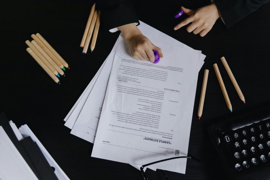 Hands working with documents and colored pencils on a black desk view.